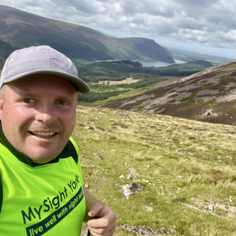 Chris is smiling at the camera during his coast to coast walk in the lowest left hand corner of the image. There are scenic views in the background.