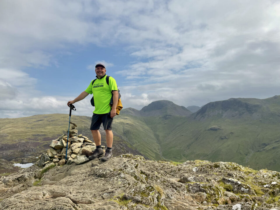 Chris is stood on top of a hill, smiling at the camera wearing a bright MySight York fundraising t-shirt.